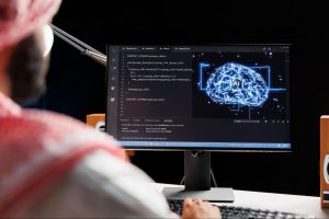 Homem usando um lenço na cabeça, sentado em frente a um monitor que exibe código de programação e uma imagem holográfica de um cérebro com o rótulo 'AI'.