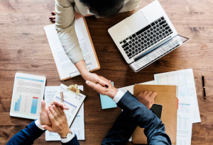 Vista aérea de três pessoas em uma reunião de negócios, apertando as mãos sobre uma mesa de madeira cheia de papéis, gráficos, relatórios e um laptop, representando uma colaboração bem-sucedida e um acordo firmado.