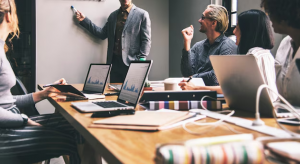 Equipe de negócios em uma sala de conferências, com laptops e cadernos sobre a mesa, assistindo a uma apresentação feita por um homem que está apontando para um quadro branco, simbolizando uma reunião de planejamento estratégico.