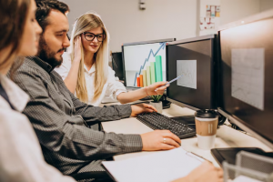 "Três pessoas trabalhando em uma mesa com vários monitores exibindo gráficos e dados, discutindo e apontando para as telas, simbolizando a análise de dados e colaboração em equipe."
