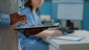 Médica utilizando um tablet enquanto outra profissional trabalha ao fundo em um consultório.